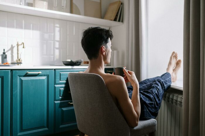 Young man sitting in kitchen chair with feet on windowsill, holding cup, reflecting on unresolved mysteries he carries.