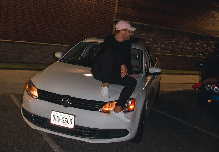 Young man in casual clothes sitting on a white car at night, reflecting the mood of revenge stories with a bitter aftertaste.