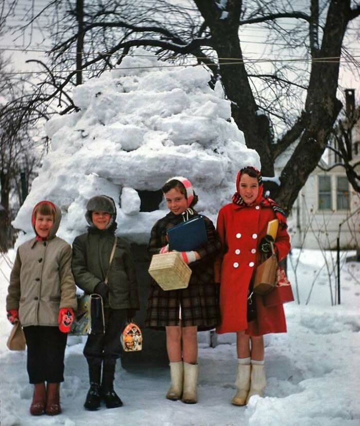 Four children dressed in winter clothes standing outside a large snow igloo in a snowy historical scene.