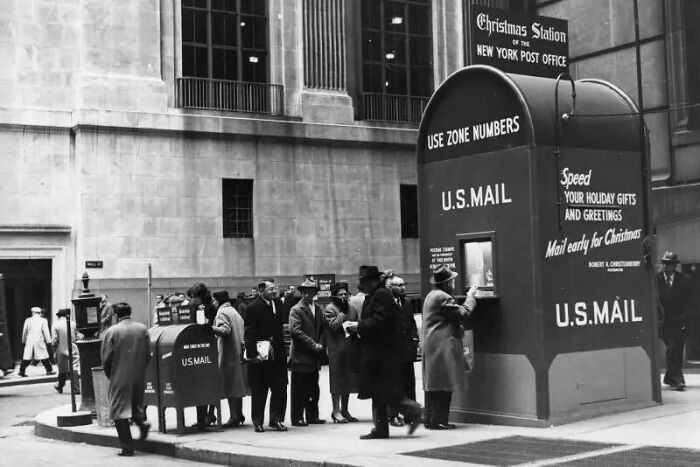 People in coats lining up at a vintage U.S. mail drop box in a historical black and white city street scene.