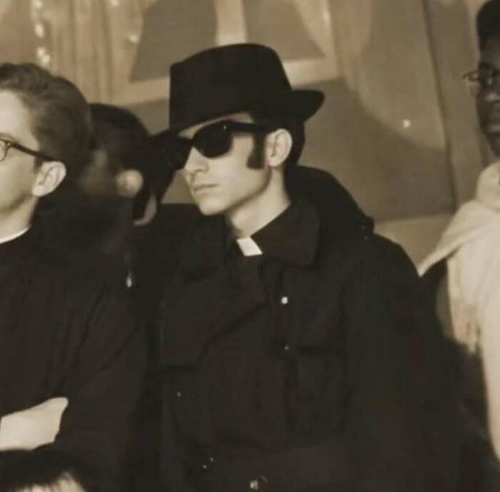 Young man wearing sunglasses and a hat, dressed as a classic priest in a historical black and white photo.