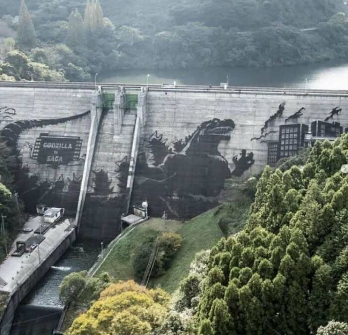 Mural of Godzilla attacking a city painted on a large dam surrounded by forest in a classic historical picture.