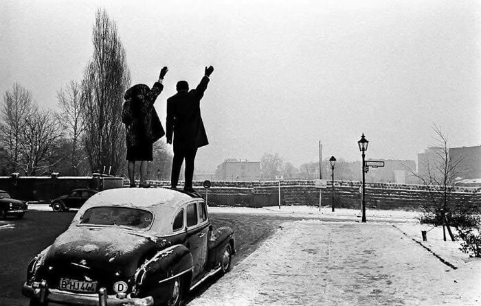 Two people standing on a snowy classic car waving goodbye in a historic black and white iconic photograph.