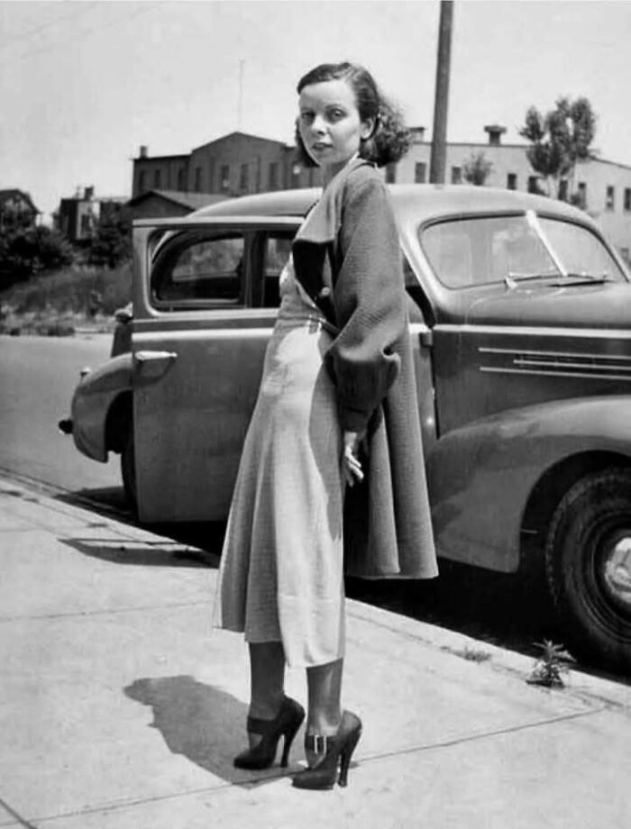 Young woman in vintage clothing standing beside a classic car in a historical black and white picture.