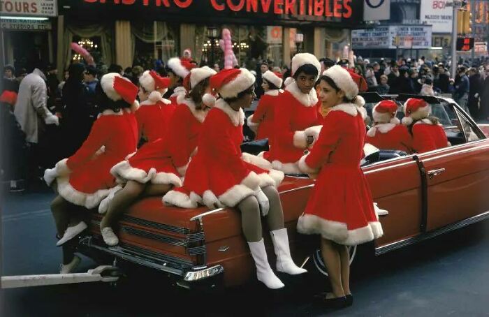 Women dressed in red and white Santa outfits sitting on a classic car during a historical parade with a large crowd in the background