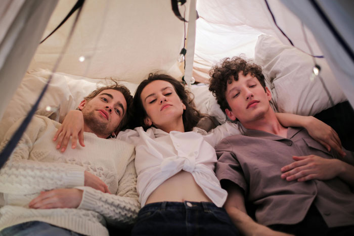 Three young adults lying closely together in a tent, illustrating polyamorous partners and family dynamics. Three young adults lying closely together in a tent, illustrating polyamorous partners and family dynamics.