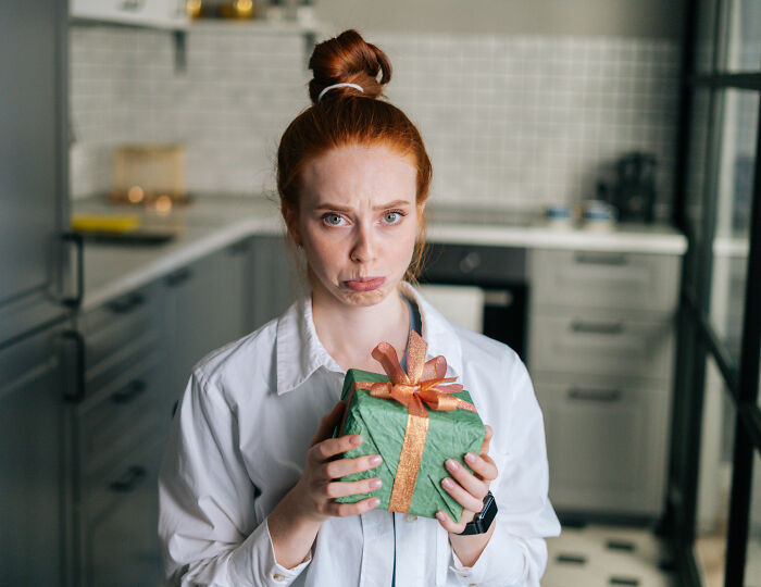 Red-haired woman holding holiday gift with a sad expression, suggesting painful truth about relationship and partner&rsquo;s gift.