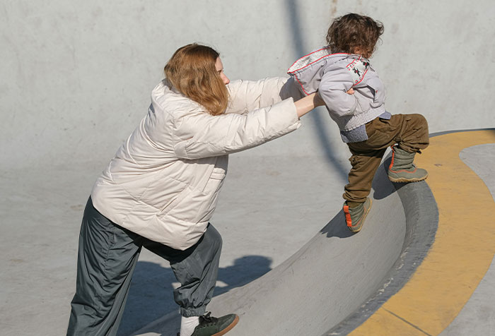 Woman supporting child climbing a curved concrete structure, illustrating strength and resilience against toxic masculinity.
