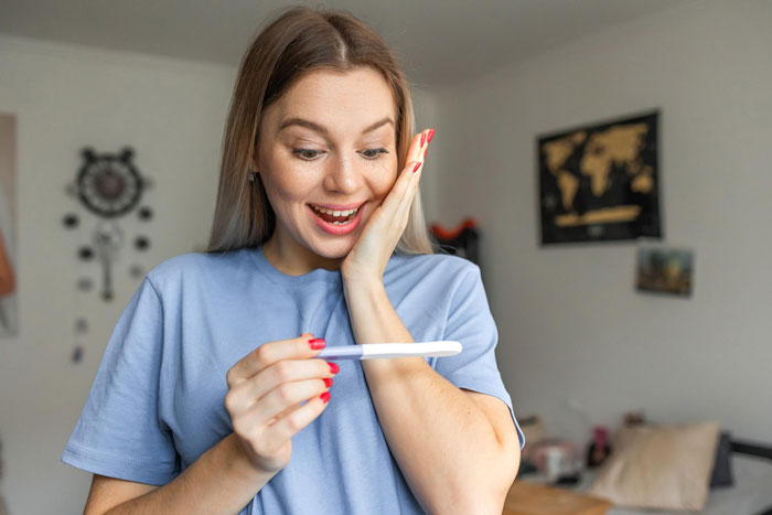 Woman in blue shirt excitedly looking at positive pregnancy test showing desire for baby before turning 30.