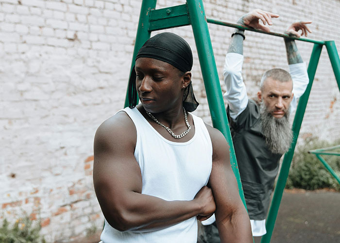 Two inmates in prison yard, one in a white tank top and durag, the other with a long beard stretching on bars, prison life misconceptions