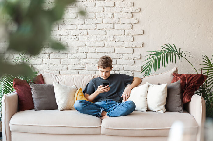Teen boy sitting cross-legged on a couch using his phone, illustrating habits affecting mental and physical health long-term.