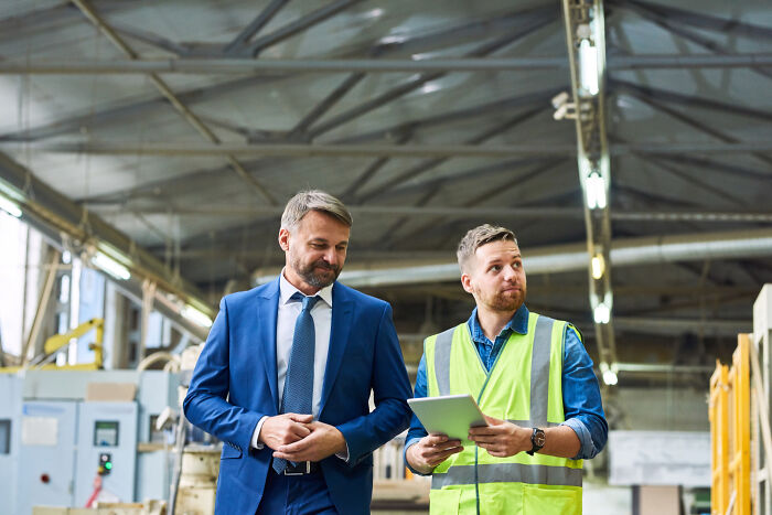Two men in a warehouse, one in a suit and the other in a safety vest, discussing quitting jobs and work experiences.