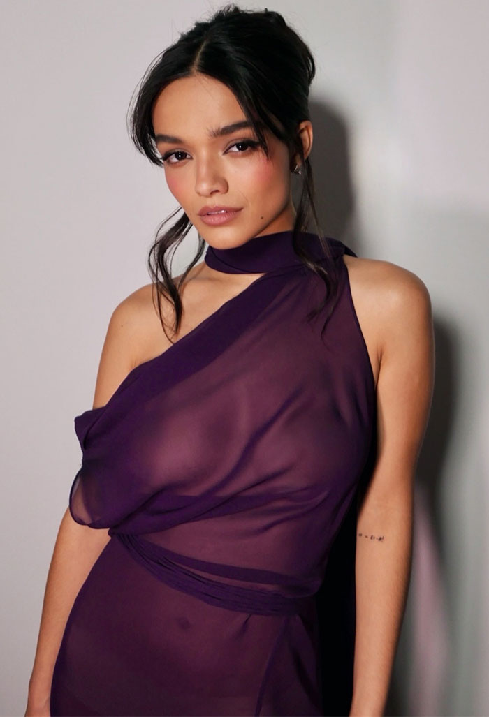 Rachel Zegler wearing a purple see-through dress with an asymmetrical design and natural makeup against a plain background. Rachel Zegler wearing a purple see-through dress with an asymmetrical design and natural makeup against a plain background.