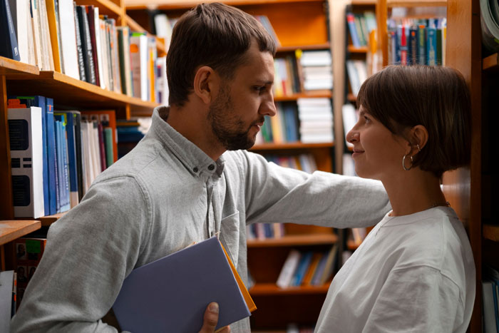 Man leaning close to younger female colleague in library aisle, she looks uneasy, suggesting inappropriate intentions suspected. Man leaning close to younger female colleague in library aisle, she looks uneasy, suggesting inappropriate intentions suspected.