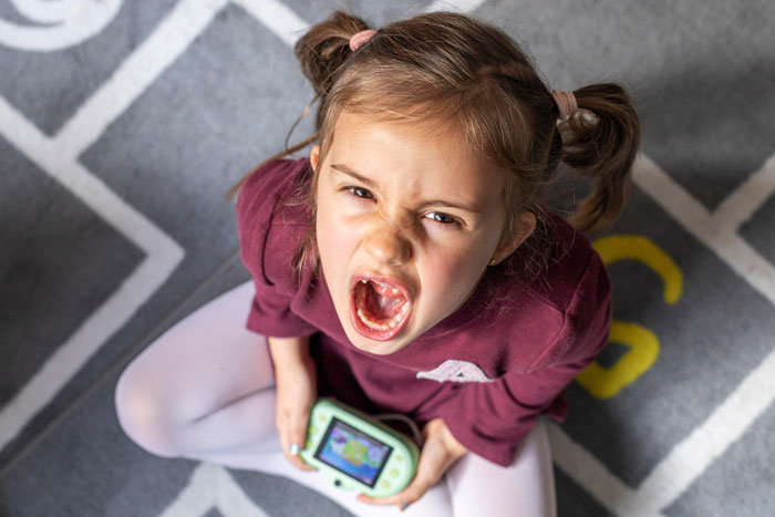 Reckless 8-year-old girl yelling while holding a handheld game console, sitting on a patterned carpet.