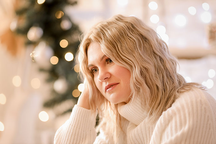 Woman in white sweater looking thoughtful near Christmas tree with lights, reflecting on hosting Christmas dinner demands.