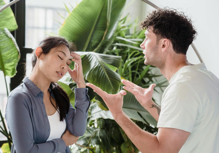 Young man arguing with woman indoors, illustrating HOA snitch conflict over grass height with tape measure and rulebooks nearby.