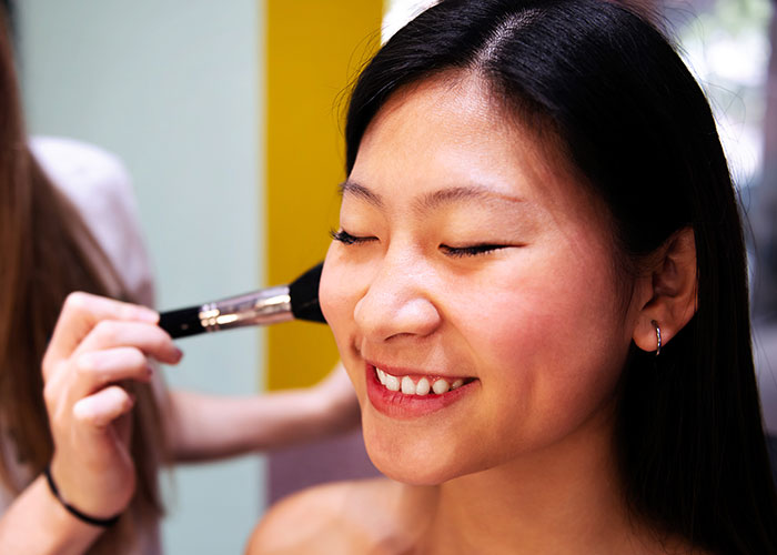 Woman smiling with eyes closed as makeup brush is applied to her cheek in a light and natural makeup setting.