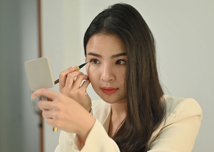Young woman applying eyeliner while holding a mirror, illustrating makeup and the no makeup look discussion online.