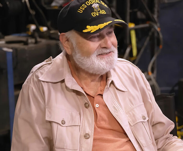 Bearded man wearing USS OORAL SEA baseball cap and beige jacket, smiling in an indoor setting discussing MAGA hypocrisy.