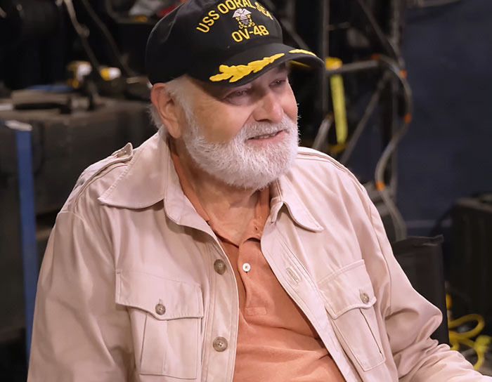 Elderly man wearing USS OORAL seal hat and beige jacket, son eyed as person of interest in LA home slaying case.