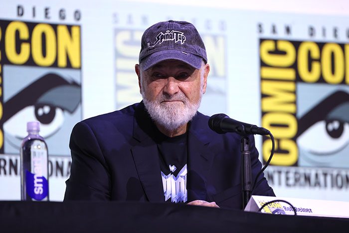 Rob Reiner at Comic-Con panel, wearing a Spinal Tap cap, seated behind a microphone with branded backdrop in the background Rob Reiner at Comic-Con panel, wearing a Spinal Tap cap, seated behind a microphone with branded backdrop in the background