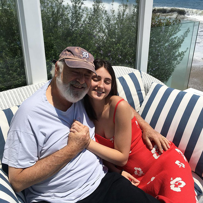 Rob Reiner’s children Jake and Romy embracing outdoors on striped cushions near greenery and ocean waves. Rob Reiner’s children Jake and Romy embracing outdoors on striped cushions near greenery and ocean waves.