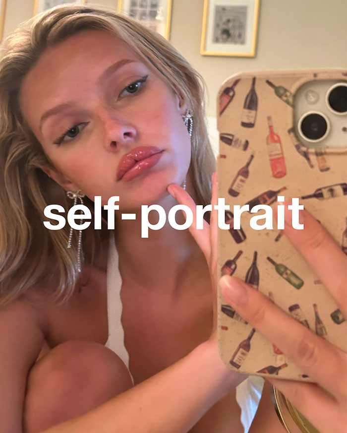 Apple Martin taking a selfie with phone case, promoting self-portrait modeling campaign, wearing earrings and white top indoors.