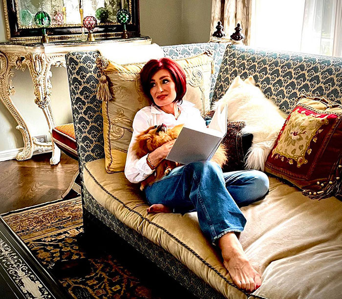 Kris Jenner sitting barefoot on a vintage sofa reading a book, showing hands without gloves after viral facelift news.