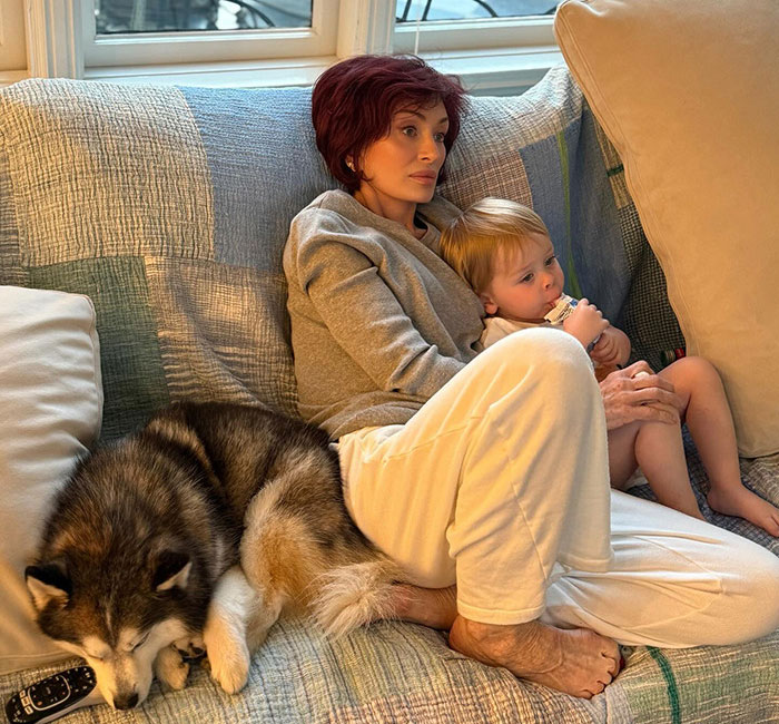 Kris Jenner sitting on a couch with a child and dog, showing hands without gloves after viral facelift news.