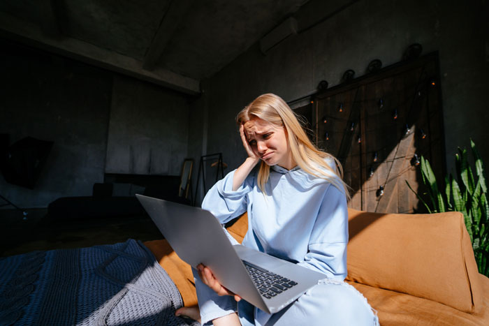 Young woman looking stressed while using a laptop indoors, planning her sil Christmas gift list on a brown sofa.