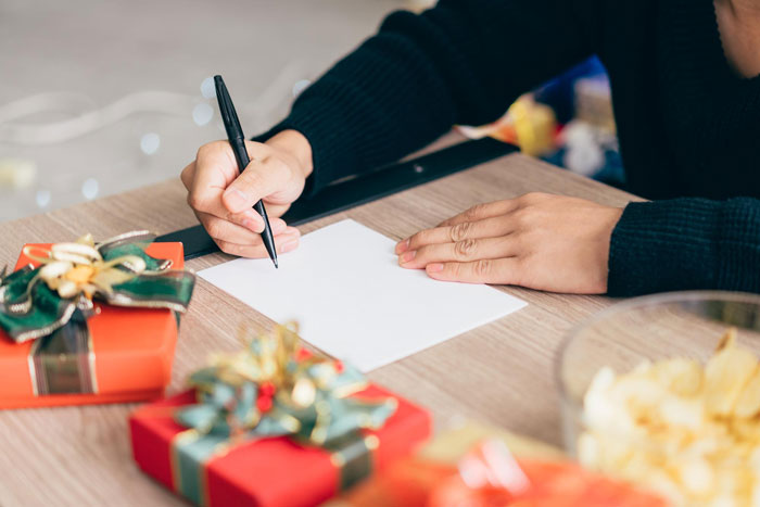 Person writing a holiday list on a blank paper surrounded by wrapped Christmas gifts and festive decorations.