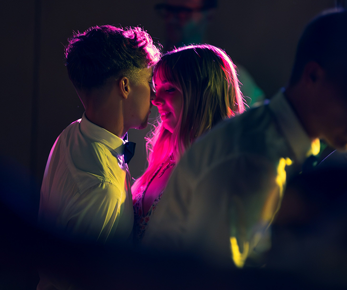 Young couple close together with the girl blushing while family members surround them at a dimly lit gathering.