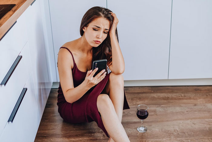 Young woman sitting on kitchen floor, looking upset while holding phone, with glass of red wine nearby, conveying tension about white baby topic.