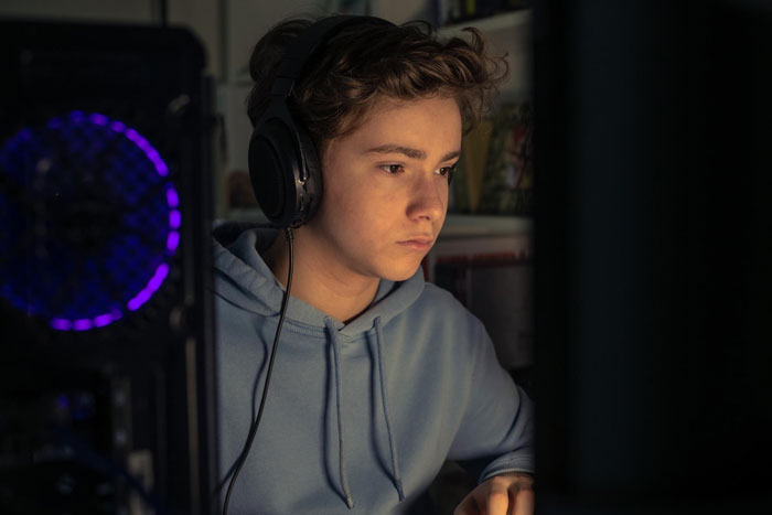 Teen boy wearing headphones focused on computer screen in dimly lit room depicting entitled behavior theme. Teen boy wearing headphones focused on computer screen in dimly lit room depicting entitled behavior theme.