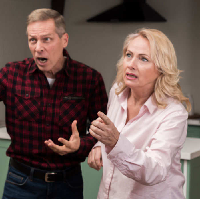 Couple arguing indoors, reacting with shock and frustration after sister&rsquo;s barefoot spiritual advisor causes comical chaos.