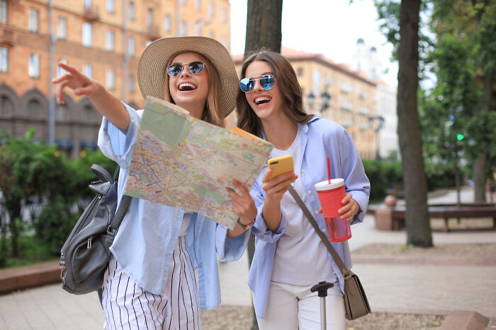 Two young women wearing sunglasses and casual clothes exploring the city with a map and smartphone, smiling and joyful.