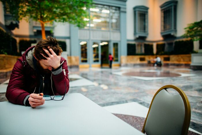 Young man looking stressed and holding glasses while sitting alone at a table, illustrating a plot twist in success stories.