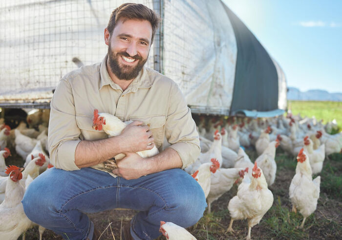 Smiling man holding a chicken on a farm surrounded by chickens, illustrating the unexpected plot twists in success stories.