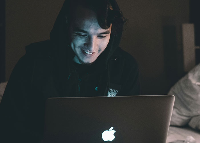 Young man in a dark hoodie smiling while using a laptop, representing diabolical revenge stories and cunning plans.