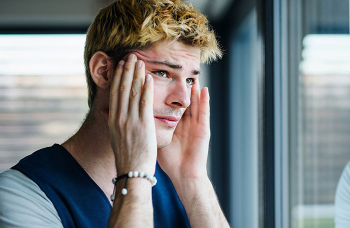 Young man holding his face looking stressed by a window, illustrating intense emotions in diabolical revenge stories.