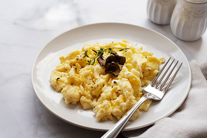Scrambled eggs on white plate with fork, garnished with herbs and truffle, showing subtle details and textures.
