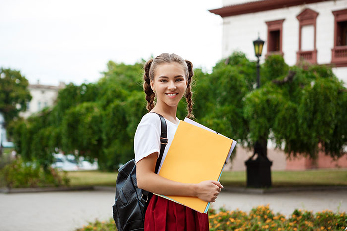 Smiling schoolgirl with backpack holding folders outside near greenery, showing no panic on her face despite serious moments