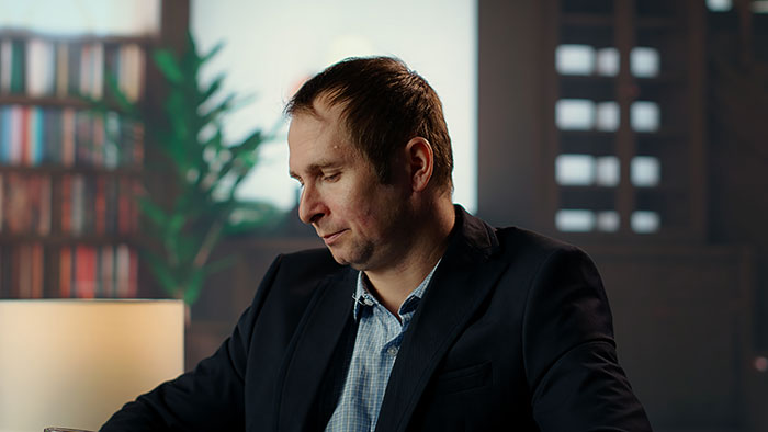 Man in a suit looking down with panic on his face in a dimly lit room, conveying serious emotions.