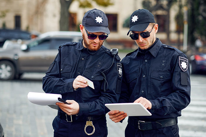Two police officers in uniform reviewing documents and a tablet while standing outdoors on a city street.