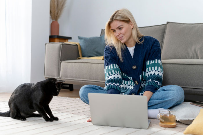 Woman sitting on the floor with laptop and black cat nearby, illustrating stealing wrong cat while paying bills concept.