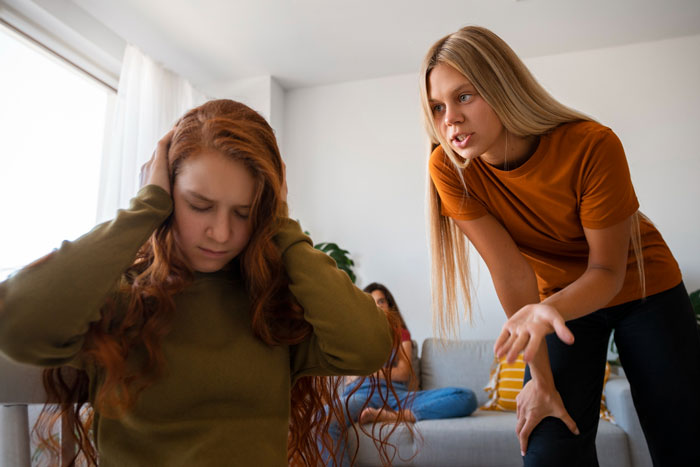 Teen girl covering ears while stepmom angrily talks, showing hostility during a tense family moment at home.