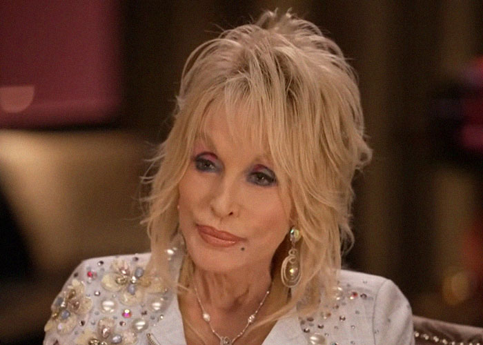 Blonde celebrity wearing jeweled jacket and large earrings, giving off elegant and confident vibes in an interview setting.