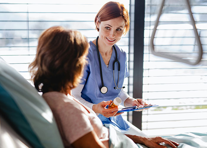 Nurse in scrubs with stethoscope talking to patient in hospital bed about post-anesthesia care and recovery.