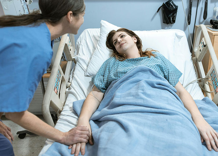 Patient in hospital bed covered with a blanket, interacting with a nurse, illustrating post-anesthesia acts in healthcare.